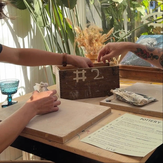 Two people reaching into a wooden box labeled '#2' on a table with plants in the background.