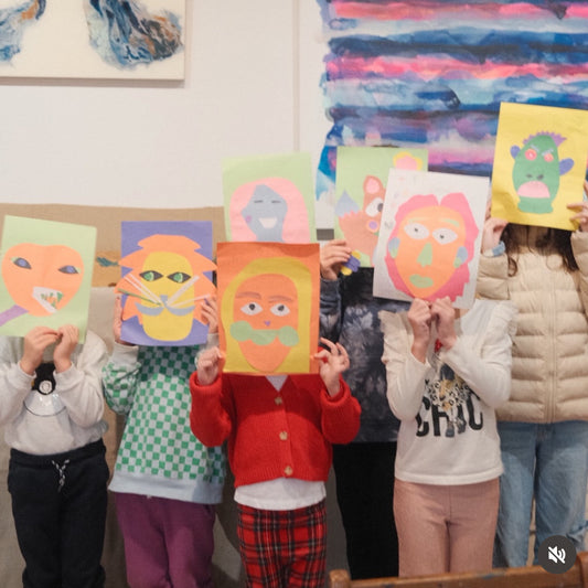 Children holding up colorful drawings of faces in front of their faces against a wall with abstract art.